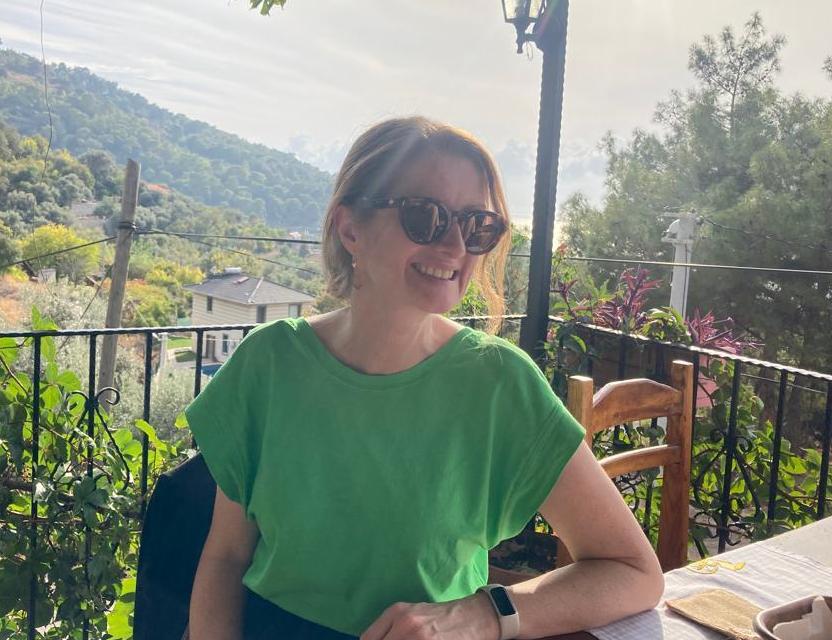 Picture of Helen: Woman on restaurant terrace with green mountains behind. Her auburn hair is tied back and she is wearing sunglasses and a green t-shirt. 